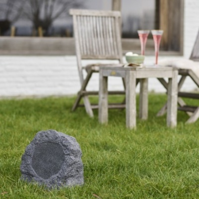 Coluna de som cinzenta em forma de pedra na relva com cadeira e mesa de madeira ao fundo