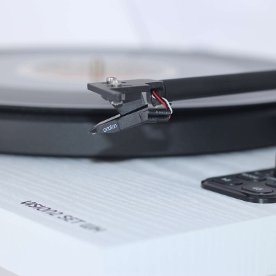 Braço de gira-discos preto com agulha Ortofon numa base branca com texto 'VISION SPIRIT' e disco de vinil cinza ao fundo