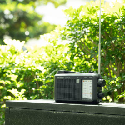 Rádio portátil preto SANGEAN sobre superfície preta com vegetação ao fundo