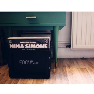 Caixa preta com discos de vinil, incluindo um da Nina Simone, junto a mesa verde e chão de madeira