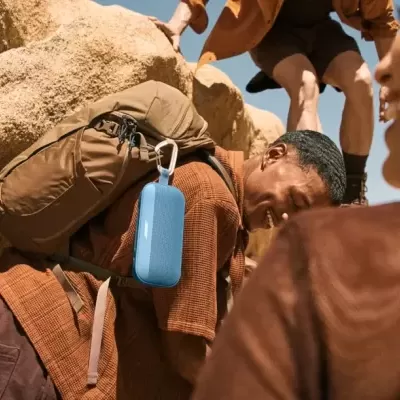 Coluna portátil azul pendurada em mochila castanha num cenário rochoso com pessoas ao redor