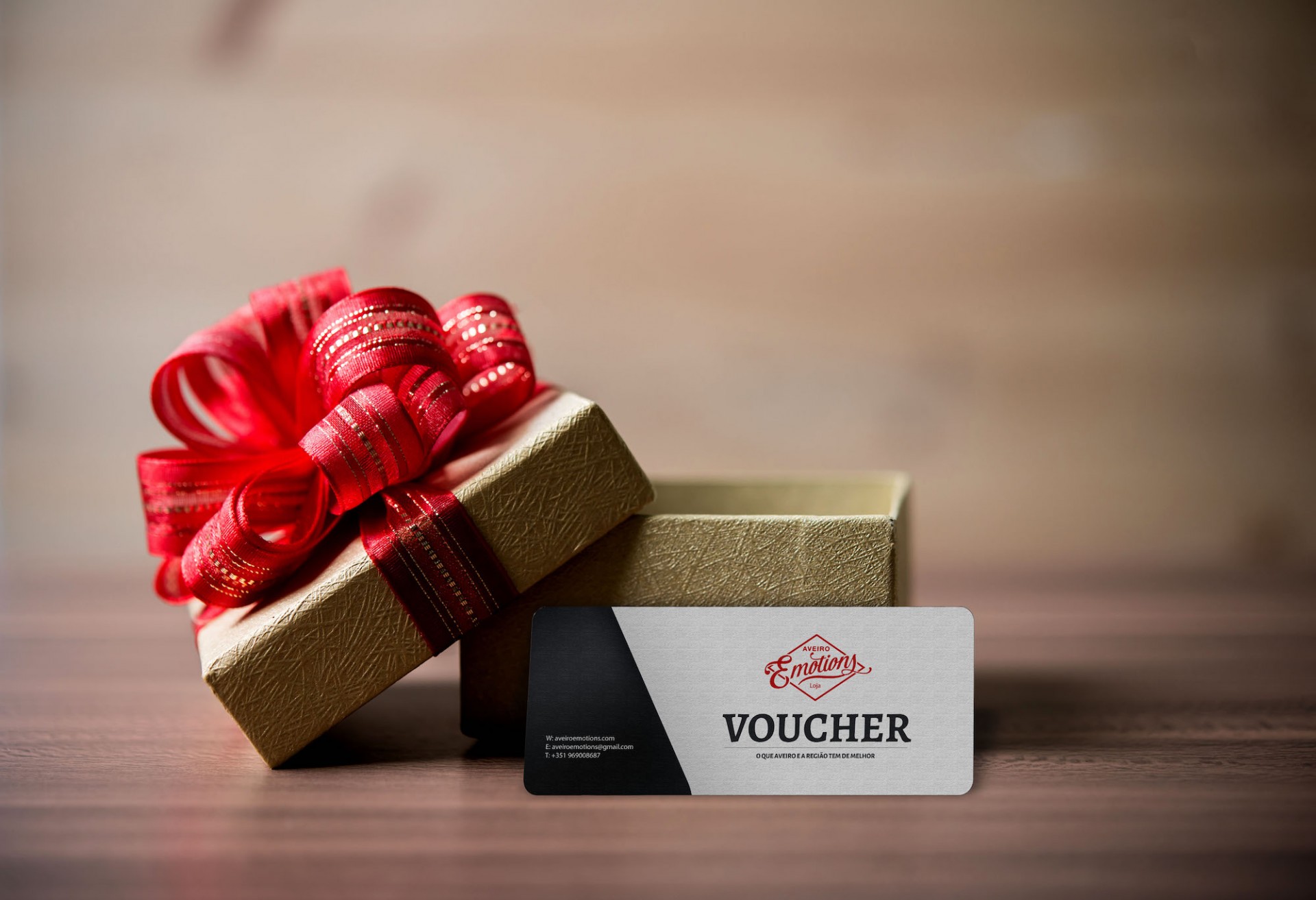 Caixa de presente dourada com fita vermelha e voucher branco e preto sobre mesa de madeira