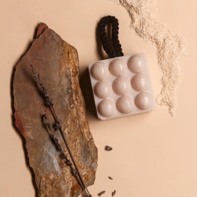 Sabonete massageador quadrado creme com nove nós e cordão preto, com pedra e raminhos secos ao lado