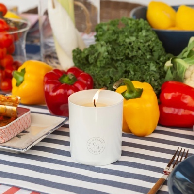 Vela branca num copo de vidro com marca CASTELBEL, rodeada por vegetais e frutas frescas sobre mesa riscada.