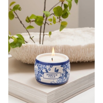 Vela em recipiente cerâmico branco e azul com texto PORTUS CALE sobre livro branco aberto com decoração natural ao fundo