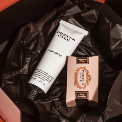 Creme de mãos e pacote rosa claro com detalhes dourados e pretos da marca PORTUS CALE dentro de caixa preta.