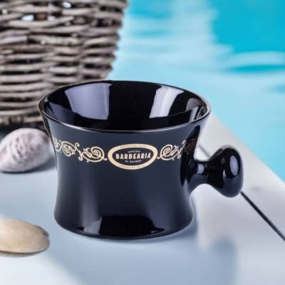 Copo de barbear preto cerâmico com decoração dourada e texto, conchas e cesto ao lado
