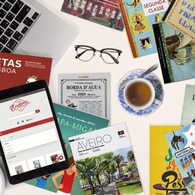 Mesa com livros portugueses, chávena de chá, laptop, tablet e óculos pretos.