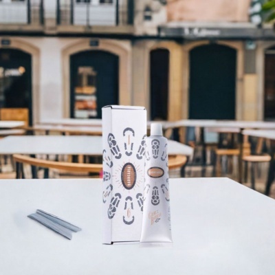 Tubo e caixa de creme branco com padrões cinzentos e texto BARBEARIA numa mesa branca num ambiente exterior.