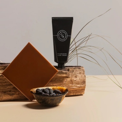 Creme para mãos perfumado em embalagem preta sobre tronco de madeira com amêndoas e placa castanha