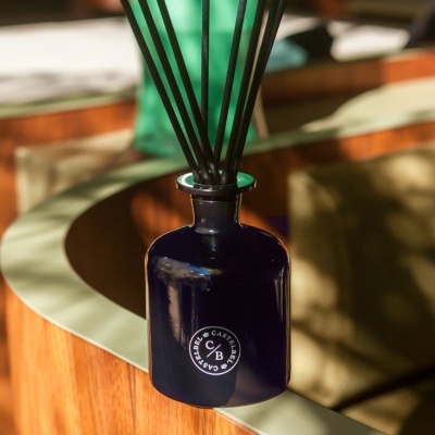 Frasco de vidro azul escuro com palitos difusores pretos e etiqueta redonda com texto CASTELBEL e C/B