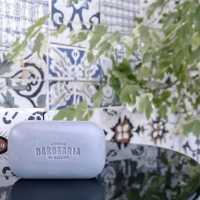 Sabonete cinza com texto ANTIGA BARBEARIA de BARRO em relevo com fundo de azulejos decorativos e folhas verdes