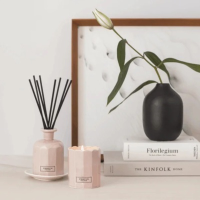 Difusor rosa, vela acesa, vaso preto com flor, livros empilhados e moldura de madeira