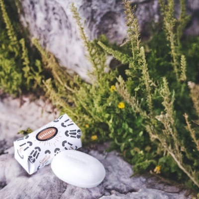 Sabonete branco oval com embalagem branca decorada junto a plantas verdes e flores amarelas numa superfície de pedra cinzenta.