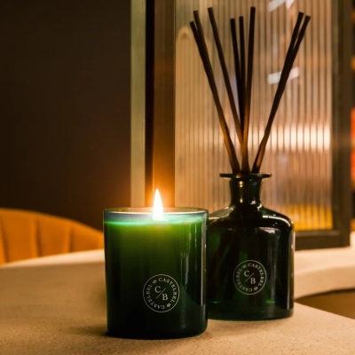 Vela perfumada verde acesa junto a difusor de ambiente verde escuro com varetas em fundo neutro