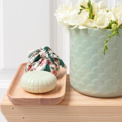 Sabonete redondo creme em bandeja de madeira com saco floral e vaso verde com flores brancas