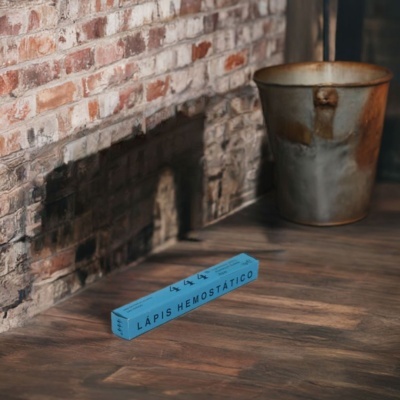 Caixa azul com texto LÁPIS HEMOSTÁTICO numa superfície de madeira junto a uma parede de tijolos e lata metálica