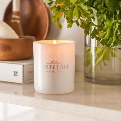 Vela branca em copo de vidro com texto ASTELBEL PORTO em superfície branca ao lado de plantas e utensílios