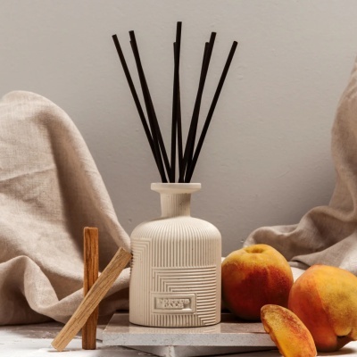 Difusor de fragrância branco com paus pretos, paus de madeira e pêssegos sobre tecido bege.