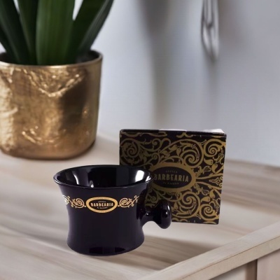 Pote preto para barbear com detalhes dourados e caixa decorativa ao lado de vaso dourado e planta
