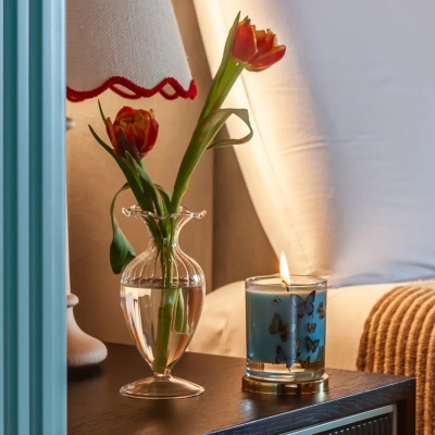 Vaso de vidro com flores e vela acesa sobre mesa de cabeceira próxima a cama.
