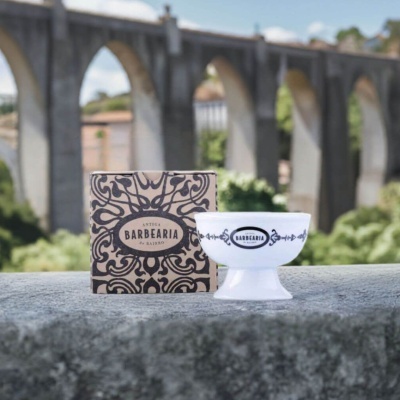 Taça de barbear branca com ornamentos e caixa de cartão com padrão preto ao lado, sobre pedra, com ponte ao fundo