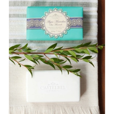 Sabonete aromático com embalagem azul-turquesa e sabonete branco CASTELBEL sobre toalha clara com ramo verde