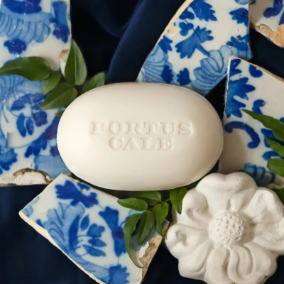 Sabonete branco oval com letras embossadas, azulejos azuis e brancos partidos e folhas verdes num fundo azul escuro.
