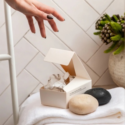 Sabonetes em forma de seixos creme e preto sobre toalhas brancas e embalagem aberta com mão feminina ao lado.