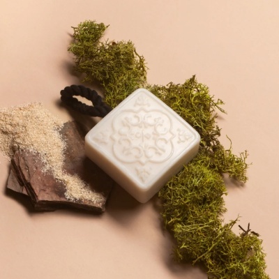 Sabonete branco quadrado com padrão decorativo e cordão preto sobre musgo e madeira