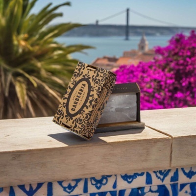 Solução de sabão em embalagem de papelão decorada com texto e flores pretas na pedra com azulejos azuis e plantas ao fundo
