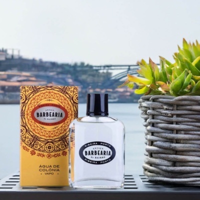 Frasco de água de colónia Antiga Barbearia de Bairro com caixa amarela e planta verde num vaso de vime cinzento ao ar livre