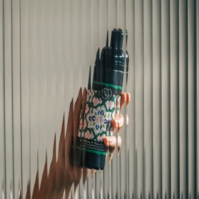 Frasco com rótulo floral colorido segurado por mão com persianas cinza ao fundo
