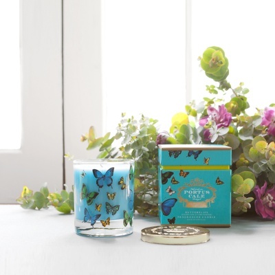Vela azul em copo de vidro com borboletas, caixa azul com borboletas e tampa dourada em superfície branca com flores.