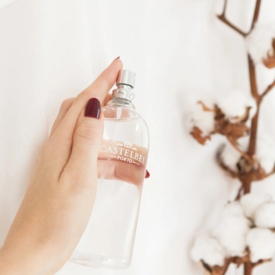 Frasco transparente com pulverizador e texto CASTELBEL PORTO segurado por mão com unhas vermelhas