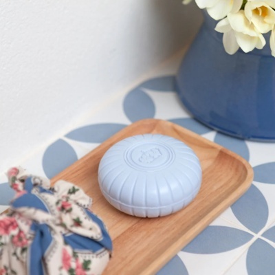 Sabonete azul redondo numa saboneteira de madeira sobre azulejos azuis e brancos com vaso e flores ao lado