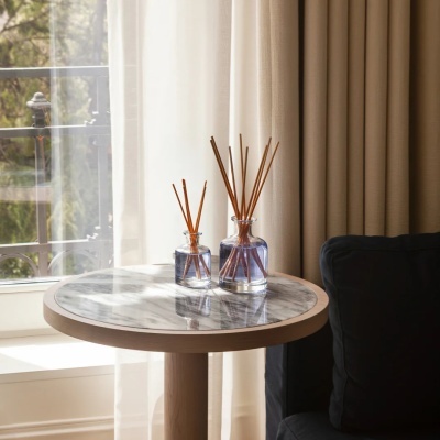 Difusores de aroma transparentes com líquido azul e varetas em mesa de madeira e mármore perto de poltrona preta e cortinas bege