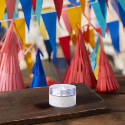 Pequeno frasco de creme com tampa prateada sobre tábua de madeira com decoração colorida ao fundo