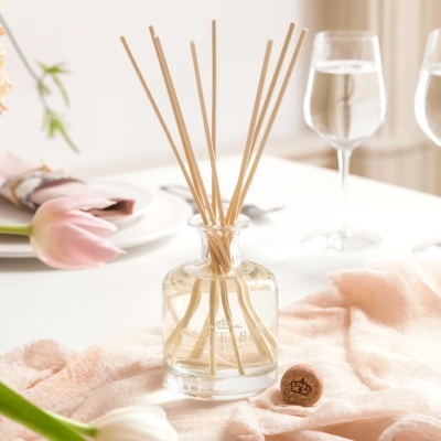 Recipiente de vidro com varetas de madeira e líquido, pano rosa, flores e copos de água.
