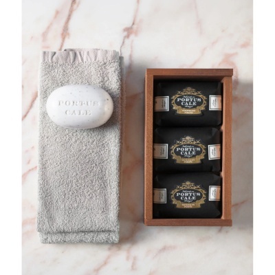 Conjunto de sabonetes Portus Cale numa caixa de madeira e sabonete branco oval com toalha cinza sobre mármore claro