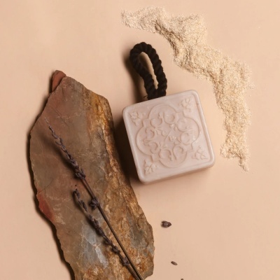 Sabonete branco com padrão e cordão preto ao lado de pedra e ramos secos de lavanda sobre fundo bege