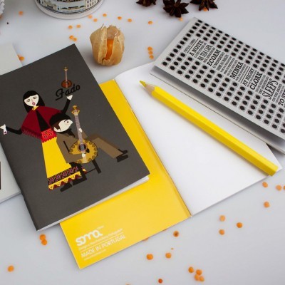 Dois cadernos com ilustrações e textos diferentes, lápis amarelo, superfície branca com pequenos grãos laranja e objeto castanho