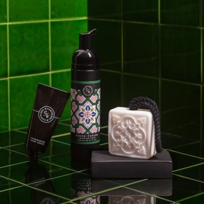 Produtos de higiene em cerâmica preta com sabonete, creme e garrafa, numa superfície com azulejos verdes.