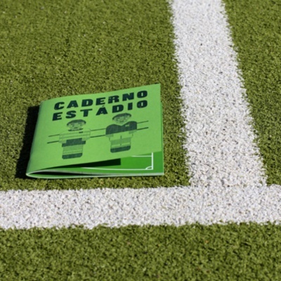 Caderno verde com capa CADERNO ESTÁDIO sobre relva sintética com linhas brancas