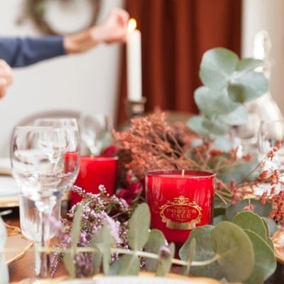 Vela vermelha com etiqueta PORTUS CALE entre flores secas e folhas numa mesa posta com copos e vela branca acesa.