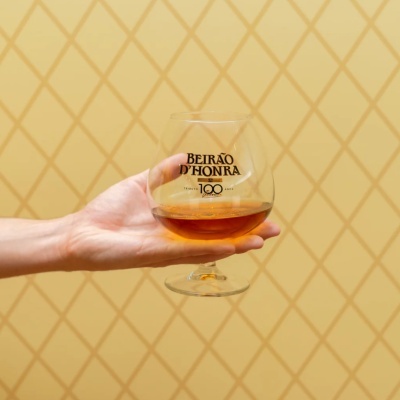 Taça de vidro com bebida âmbar e texto BEIRÃO DHONRA segurada por uma mão sobre fundo amarelo com padrão.