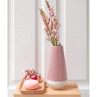 Vaso cerâmico rosa com flores artificiais, sabonete rosa oval e saco de tecido vermelho em bandeja de madeira sobre mesa