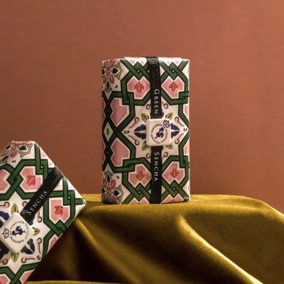 Embalagens retangulares decoradas com padrão geométrico e fita preta com texto, sobre pano dourado