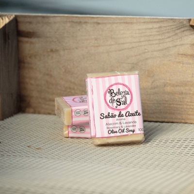 Sabonetes de azeite com embalagem rosa com riscas brancas e texto preto colocados numa superfície de malha numa caixa de madeira.