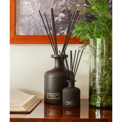 Dois difusores de aroma em vidro fosco preto com rótulos PORTUS CALE sobre mesa de madeira com livro e vaso com folhagem.